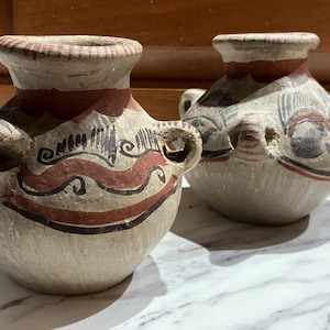 May include: Two small, antique ceramic vases with a cream-colored base and red and black painted designs. Each vase has a rounded body, a narrow neck, and two small handles. The vases are displayed on a white marble surface.