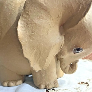Peut inclure: Une figurine d'éléphant de couleur beige avec une surface texturée et mate. L'éléphant est représenté dans une position tournée vers le bas avec de grandes oreilles proéminentes, une trompe courbée et un œil visible avec une pupille sombre.