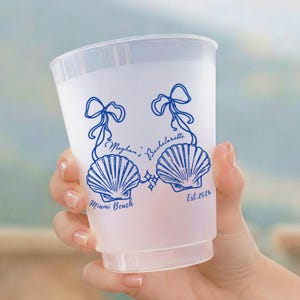 May include: A translucent frosted plastic cup featuring a blue graphic print of two seashells tied with ribbons. The text reads Meghan's Bachelorette, Miami Beach, and Est. 2026. The design includes small star accents between the two seashells.