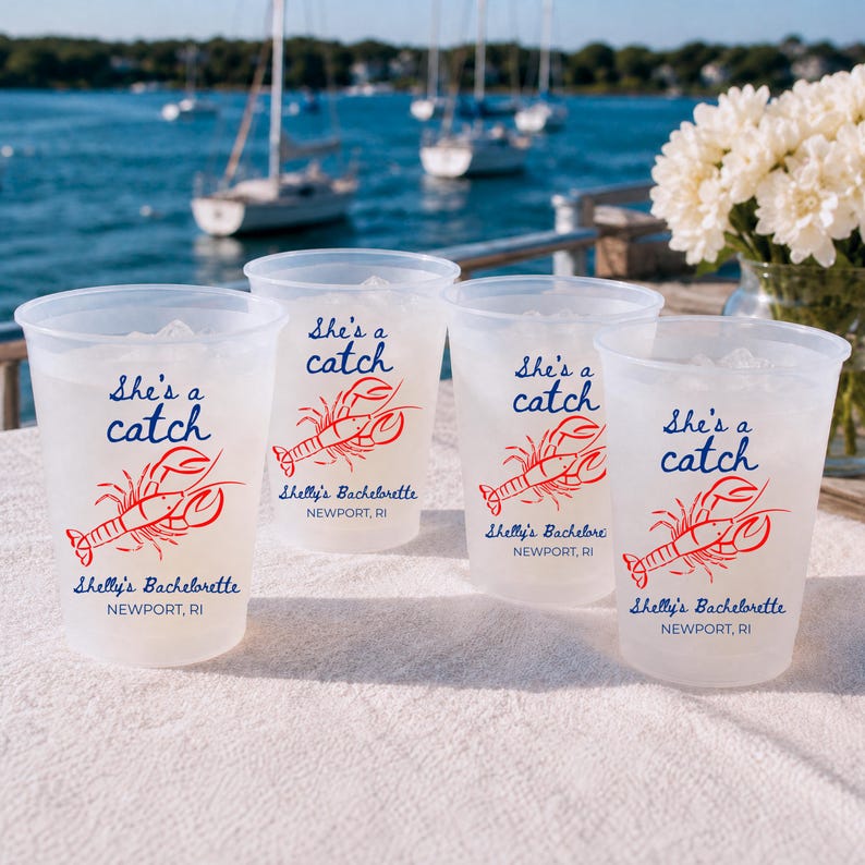 May include: Four translucent plastic cups filled with ice, each featuring a red lobster illustration and blue text that reads She's a catch, Shelly's Bachelorette, and NEWPORT, RI. The cups are arranged on a white surface with a harbor view.