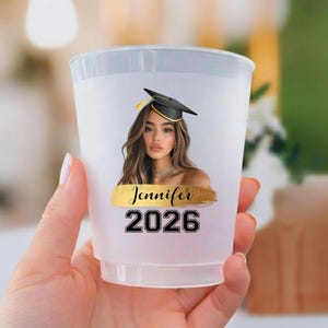 May include: A translucent plastic cup featuring a printed graphic of a person wearing a black graduation cap. Below the image is a gold brushstroke design with the name Jennifer printed in black script, followed by the graduation year 2026.