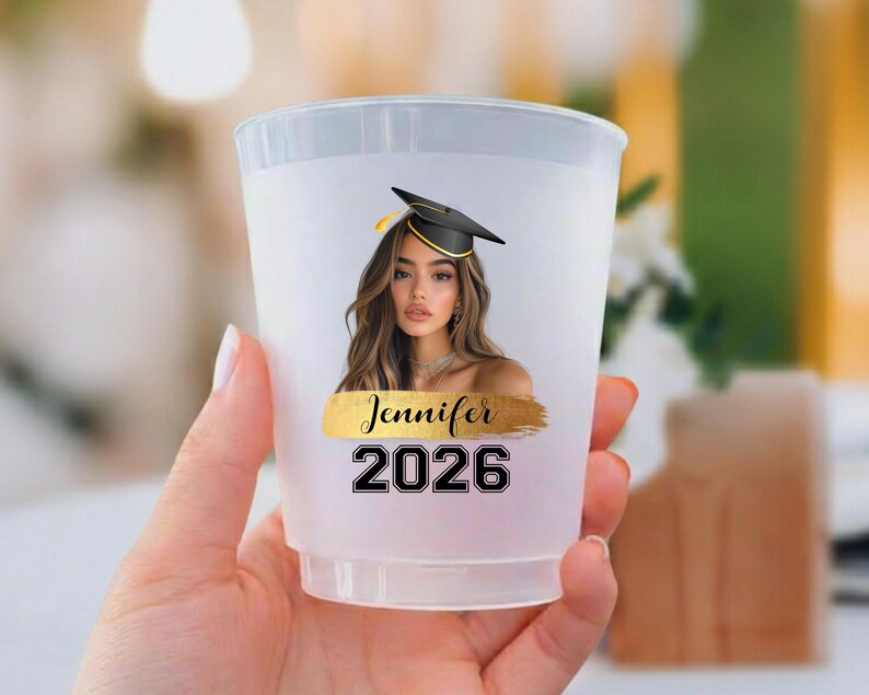 May include: A translucent plastic cup featuring a printed graphic of a person wearing a black graduation cap. Below the image is a gold brushstroke design with the name Jennifer printed in black script, followed by the graduation year 2026.