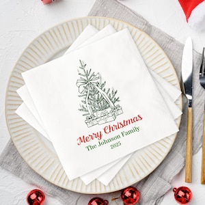 May include: White square paper napkins with a Christmas tree design, a bow, and gift boxes. The napkins have the text "Merry Christmas" in red and "The Johnson Family 2025" in green. The napkins are on a white plate with a knife and fork.