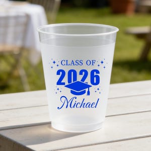 May include: A frosted translucent plastic cup featuring blue printed text that reads "CLASS OF 2026" above a graphic of a graduation cap and the name "Michael" in a cursive script, accented by small star shapes surrounding the central design.