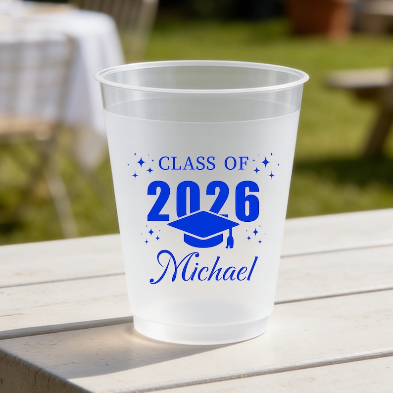 May include: A frosted translucent plastic cup featuring blue printed text that reads "CLASS OF 2026" above a graphic of a graduation cap and the name "Michael" in a cursive script, accented by small star shapes surrounding the central design.