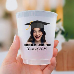 May include: A translucent plastic party cup featuring a printed graphic of a person wearing a black graduation cap. The text reads CONGRATS JENNIFER! Class of 2026 in black lettering. The cup has a smooth, tapered cylindrical shape and a rim.