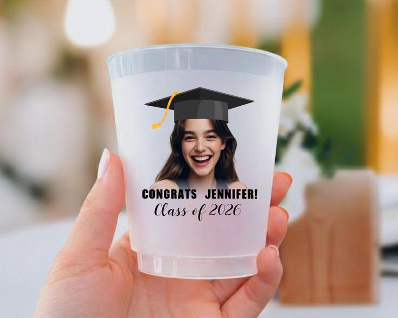 May include: A translucent plastic party cup featuring a printed graphic of a person wearing a black graduation cap. The text reads CONGRATS JENNIFER! Class of 2026 in black lettering. The cup has a smooth, tapered cylindrical shape and a rim.