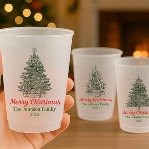 May include: Three frosted plastic cups featuring Christmas tree illustrations and the text "Merry Christmas The Johnson Family 2025" in red and green. One cup is held, the others are on a surface.