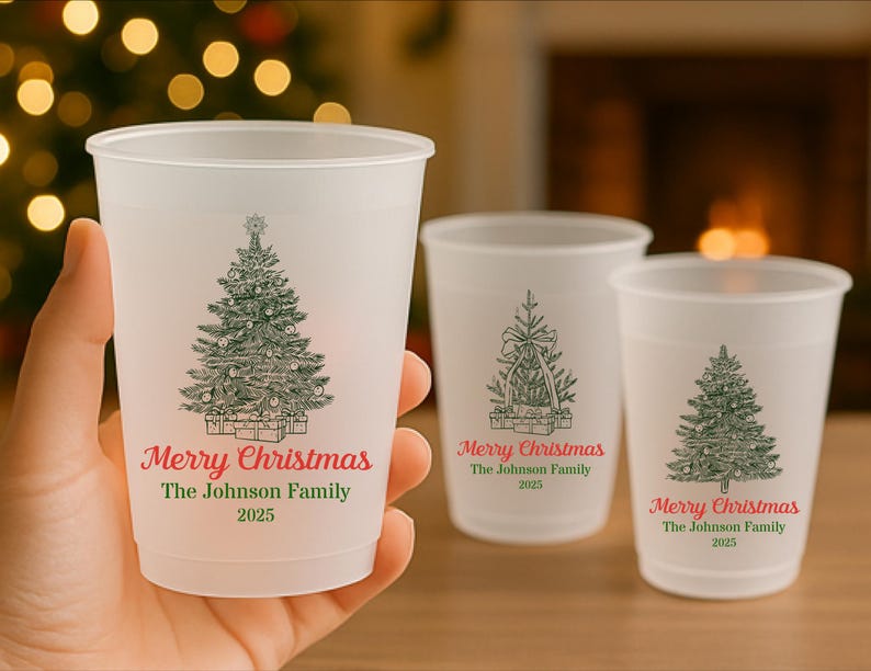 May include: Three frosted plastic cups featuring Christmas tree illustrations and the text "Merry Christmas The Johnson Family 2025" in red and green. One cup is held, the others are on a surface.