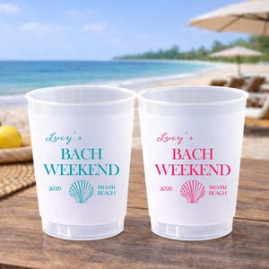 May include: Two translucent plastic cups featuring the text Lucy's BACH WEEKEND 2026 MIAMI BEACH and a seashell graphic. One cup has teal text and graphics, while the other cup features identical text and a seashell graphic in hot pink.