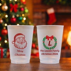 May include: Two frosted plastic cups. The left cup features a red Santa Claus graphic with the text Merry Christmas. The right cup displays a green and red wreath with a red letter L in the center and the text The Lawson Family Christmas 2026.