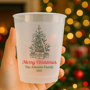 May include: A frosted plastic cup with a green Christmas tree design, bow, and presents. The text "Merry Christmas" and "The Johnson Family 2025" are printed in red and green. The cup is held in a hand.