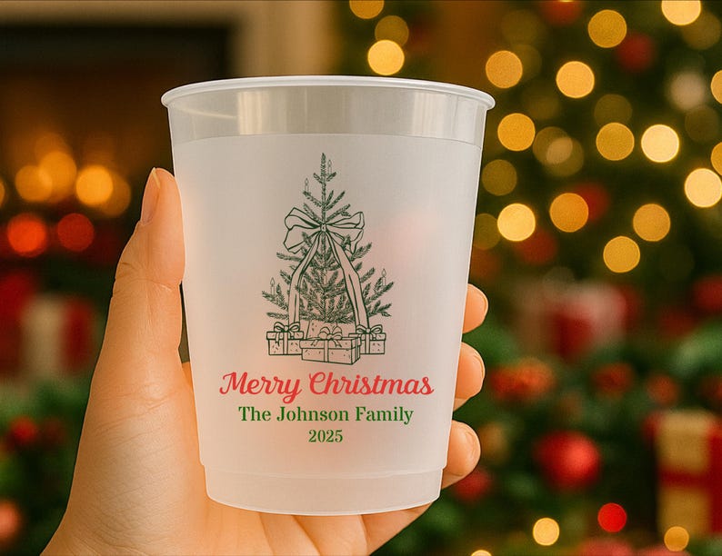 May include: A frosted plastic cup with a green Christmas tree design, bow, and presents. The text "Merry Christmas" and "The Johnson Family 2025" are printed in red and green. The cup is held in a hand.
