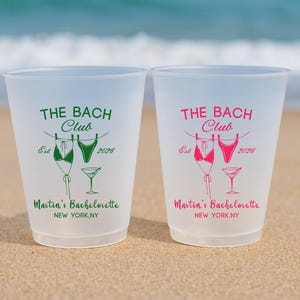 Może przedstawiać: Dwa kubki z matowego plastiku z napisem THE BACH CLUB, Est 2026, Martin's Bachelorette oraz NEW YORK, NY. Jeden kubek ma zielony nadruk, drugi r&oacute;żowy. Oba przedstawiają grafikę dw&oacute;ch bikini na sznurku do bielizny oraz kieliszek do martini.