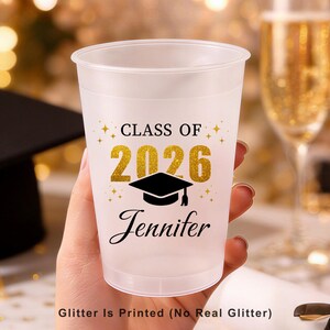 May include: A frosted plastic cup featuring the black text "CLASS OF" above the gold-coloured printed numbers "2026" and a black graduation cap icon. Below the icon, the name "Jennifer" is printed in black script. Text reads "Glitter Is Printed (No Real Glitter)".
