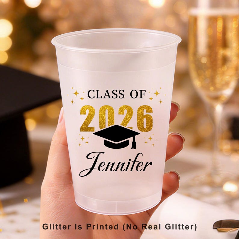 May include: A frosted plastic cup featuring the black text "CLASS OF" above the gold-coloured printed numbers "2026" and a black graduation cap icon. Below the icon, the name "Jennifer" is printed in black script. Text reads "Glitter Is Printed (No Real Glitter)".