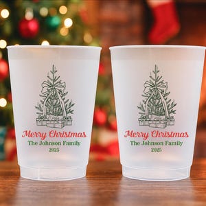 May include: Two frosted plastic cups featuring a Christmas tree design and the words "Merry Christmas" in red script. Below the text is "The Johnson Family 2025" in green. The cups are on a wooden surface with a Christmas tree and fireplace in the background.
