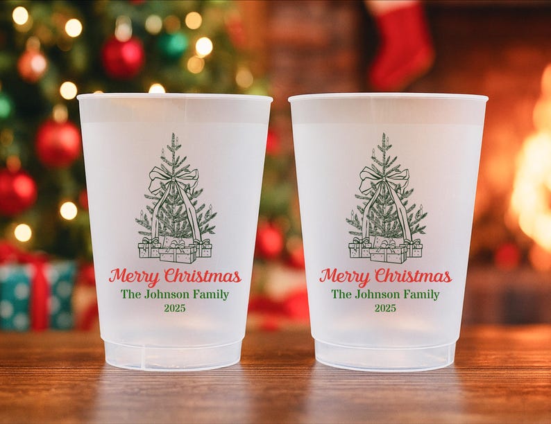 May include: Two frosted plastic cups featuring a Christmas tree design and the words "Merry Christmas" in red script. Below the text is "The Johnson Family 2025" in green. The cups are on a wooden surface with a Christmas tree and fireplace in the background.
