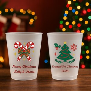 May include: Two frosted plastic cups. The left cup features red and white candy canes, a green plaid bow, and the text "Merry Christmas Ketty & James". The right cup displays a green Christmas tree, holly, and "Engaged For Christmas 2026".