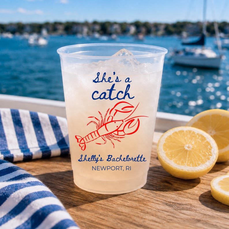 May include: A clear plastic cup filled with ice, featuring a red lobster graphic and blue text that reads She's a catch, Shelly's Bachelorette, and NEWPORT, RI. The cup is positioned on a wooden surface next to sliced yellow lemons.