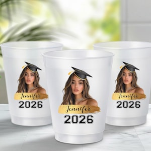 May include: Three translucent white plastic cups featuring a printed graphic of a person wearing a black graduation cap. Below the image is a gold brushstroke design with the name Jennifer in black script and the year 2026 in bold black font.