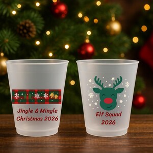 May include: Two frosted plastic cups featuring Christmas designs. One cup displays a red and green plaid pattern with the text Jingle & Mingle Christmas 2026. The other cup features a green reindeer graphic with the text Elf Squad 2026.