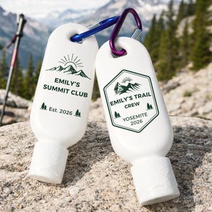 May include: Two white, oval-shaped sunscreen bottles with green and black text and graphics, attached to carabiners. One bottle reads "Emily's Summit Club Est. 2026." The other reads "Emily's Trail Crew Yosemite 2026."