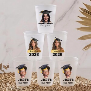 May include: A stack of six frosted plastic cups featuring personalized graduation designs. The top cup reads CONGRATS JENNIFER! Class of 2026. The middle cups feature a photo of Jennifer with the name Jennifer and 2026. The bottom cups show Jacob's face.