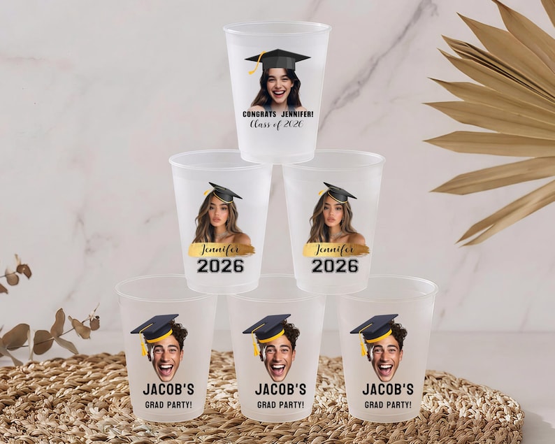 May include: A stack of six frosted plastic cups featuring personalized graduation designs. The top cup reads CONGRATS JENNIFER! Class of 2026. The middle cups feature a photo of Jennifer with the name Jennifer and 2026. The bottom cups show Jacob's face.