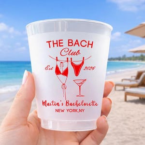 Może przedstawiać: P&oacute;łprzezroczysty plastikowy kubek z czerwonym nadrukiem. Projekt zawiera napisy THE BACH CLUB, Est 2026, Martin's Bachelorette oraz NEW YORK, NY, a także ilustracje przedstawiające bikini, str&oacute;j kąpielowy i kieliszek do martini.