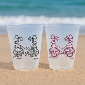 May include: Two frosted plastic cups featuring a black and a maroon graphic of two seashells tied with ribbons. The text reads Meghan's Bachelorette, Miami Beach, and Est. 2026. The cups are translucent and have a simple, cylindrical shape.