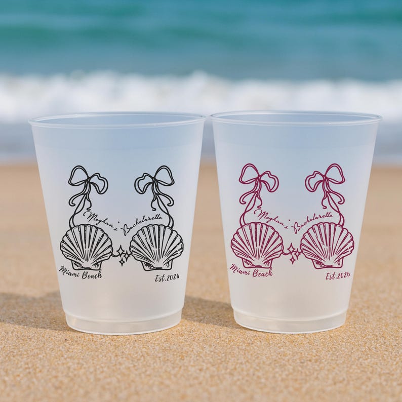 May include: Two frosted plastic cups featuring a black and a maroon graphic of two seashells tied with ribbons. The text reads Meghan's Bachelorette, Miami Beach, and Est. 2026. The cups are translucent and have a simple, cylindrical shape.