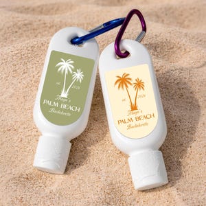 May include: Two white sunscreen bottles with blue and purple carabiner clips. Each bottle has a label with a palm tree design and the text "Maya's PALM BEACH Bachelorette". The bottles are resting on sand.