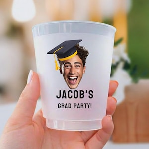 May include: A translucent plastic party cup featuring a printed graphic of a person wearing a black graduation cap with a yellow tassel. Below the face, the text reads JACOB'S GRAD PARTY! in bold black capital letters on the front surface.