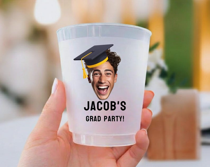 May include: A translucent plastic party cup featuring a printed graphic of a person wearing a black graduation cap with a yellow tassel. Below the face, the text reads JACOB'S GRAD PARTY! in bold black capital letters on the front surface.