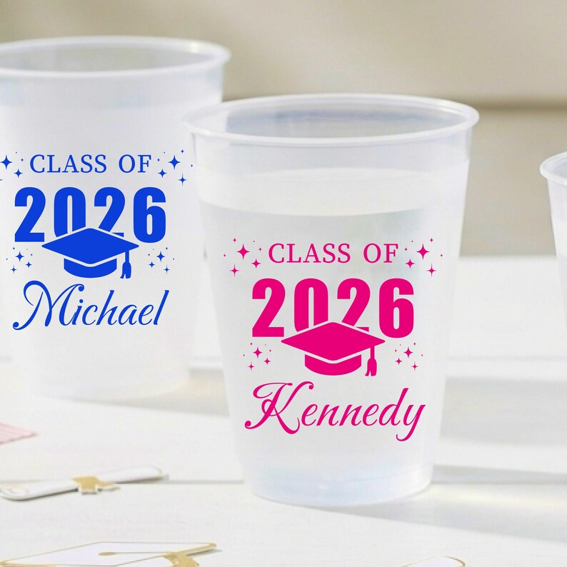 May include: Two translucent plastic cups featuring graduation-themed designs. One cup displays blue text reading "CLASS OF 2026" above a blue graduation cap and the name "Michael". The other cup features pink text and a pink graduation cap.