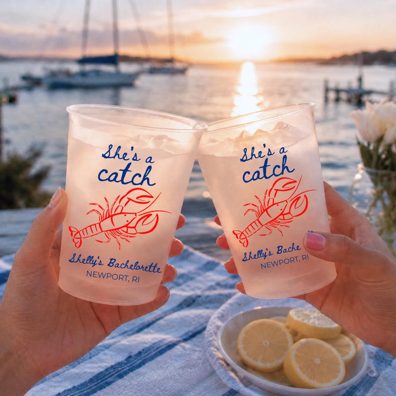 May include: Two clear plastic cups filled with ice and a light-colored beverage. Each cup features a red lobster graphic and blue text that reads "She's a catch," "Shelly's Bachelorette," and "NEWPORT, RI" printed on the front surface.