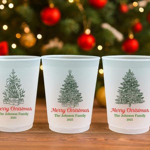 May include: Three white plastic cups with a Christmas tree design and the text "Merry Christmas The Johnson Family 2025". The cups are on a wooden surface with a blurred Christmas tree background.