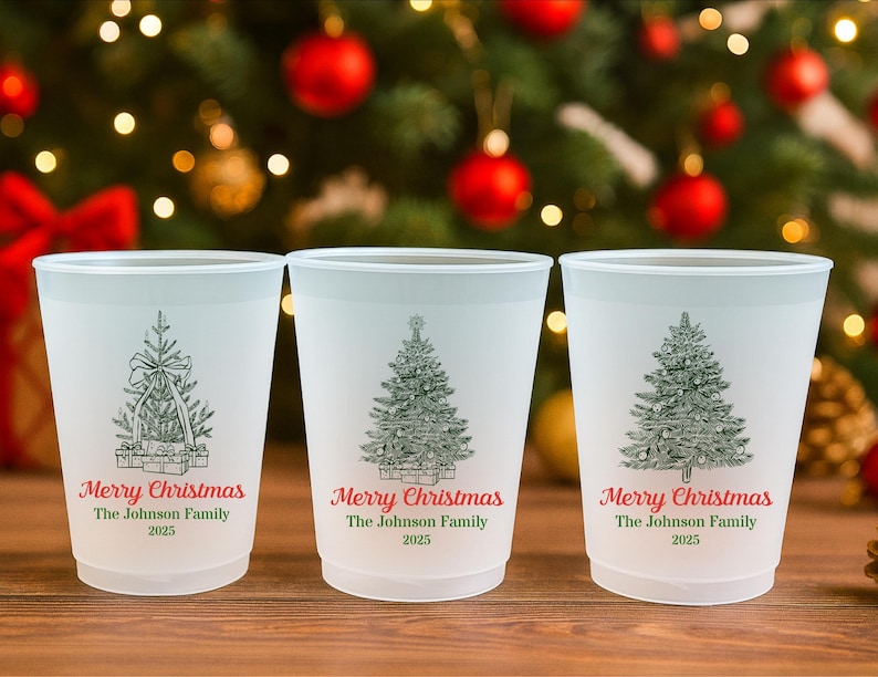 May include: Three white plastic cups with a Christmas tree design and the text "Merry Christmas The Johnson Family 2025". The cups are on a wooden surface with a blurred Christmas tree background.