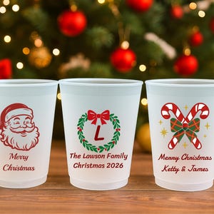 May include: Three frosted plastic cups featuring Christmas designs. The first shows a red Santa face with "Merry Christmas". The second has a green wreath with a red bow, "L", "The Lawson Family", and "Christmas 2026". The third has candy canes, "Merry Christmas", and "Ketty & James".