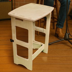 May include: A light-colored wooden step stool with a rectangular top and cut-out sides. The stool has a handle cut into the top and a black hinge on the side. The stool is approximately 18 inches tall.