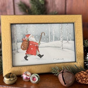 K&ouml;nnte beinhalten: Gerahmtes Aquarellgem&auml;lde von Santa Claus, der durch einen verschneiten Wald geht und einen Geschenkesack und einen Stock tr&auml;gt. Das Gem&auml;lde befindet sich in einem goldenen Rahmen. Die Signatur des K&uuml;nstlers, "K.Schmutz", ist sichtbar. Weihnachtsdekoration.
