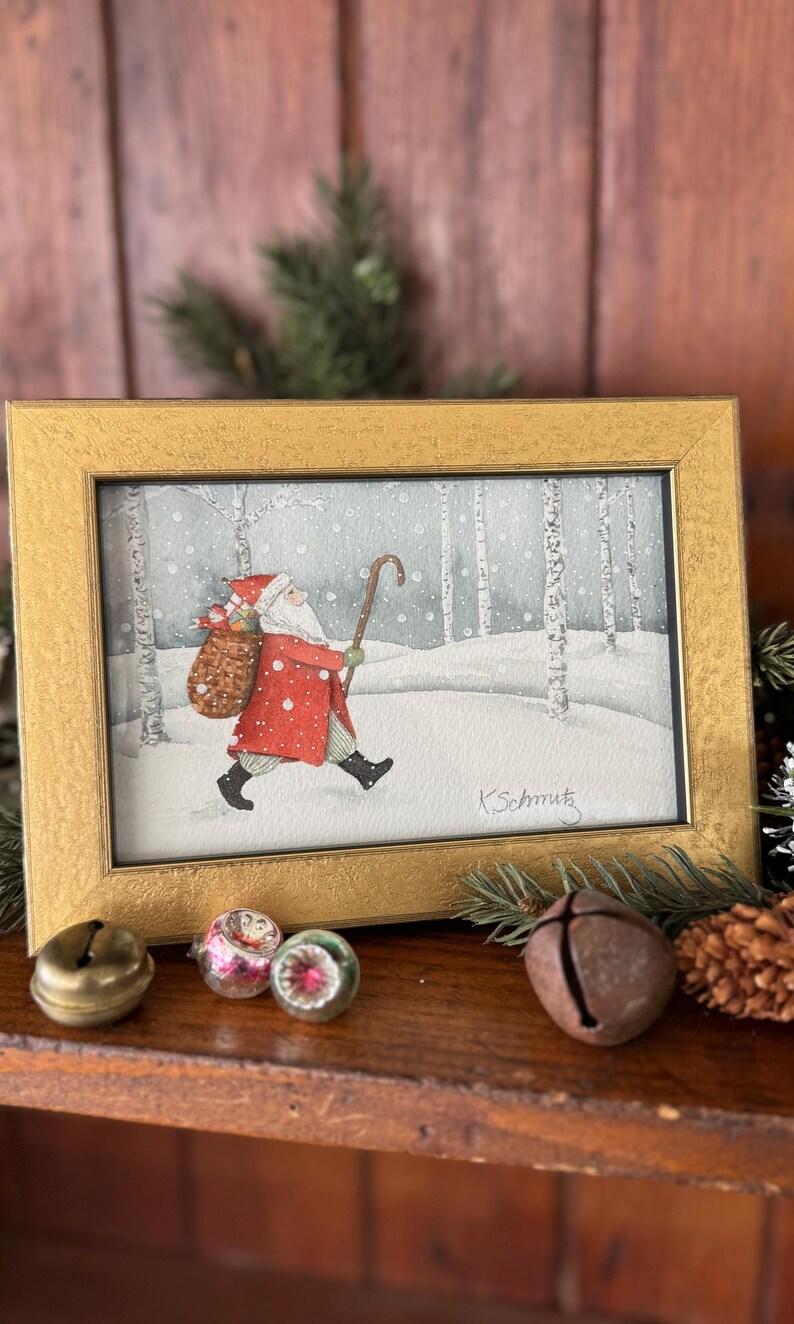 K&ouml;nnte beinhalten: Gerahmtes Aquarellgem&auml;lde von Santa Claus, der durch einen verschneiten Wald geht und einen Geschenkesack und einen Stock tr&auml;gt. Das Gem&auml;lde befindet sich in einem goldenen Rahmen. Die Signatur des K&uuml;nstlers, "K.Schmutz", ist sichtbar. Weihnachtsdekoration.