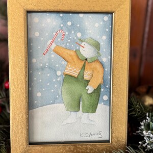K&ouml;nnte beinhalten: Aquarellmalerei eines Schneemanns in einem goldenen Rahmen. Der Schneemann tr&auml;gt einen gr&uuml;nen Hut, eine Latzhose und einen orangefarbenen Pullover und h&auml;lt eine Zuckerstange. Der Hintergrund ist eine verschneite blaue Szene. Das Gem&auml;lde ist signiert "K. Schmitz".