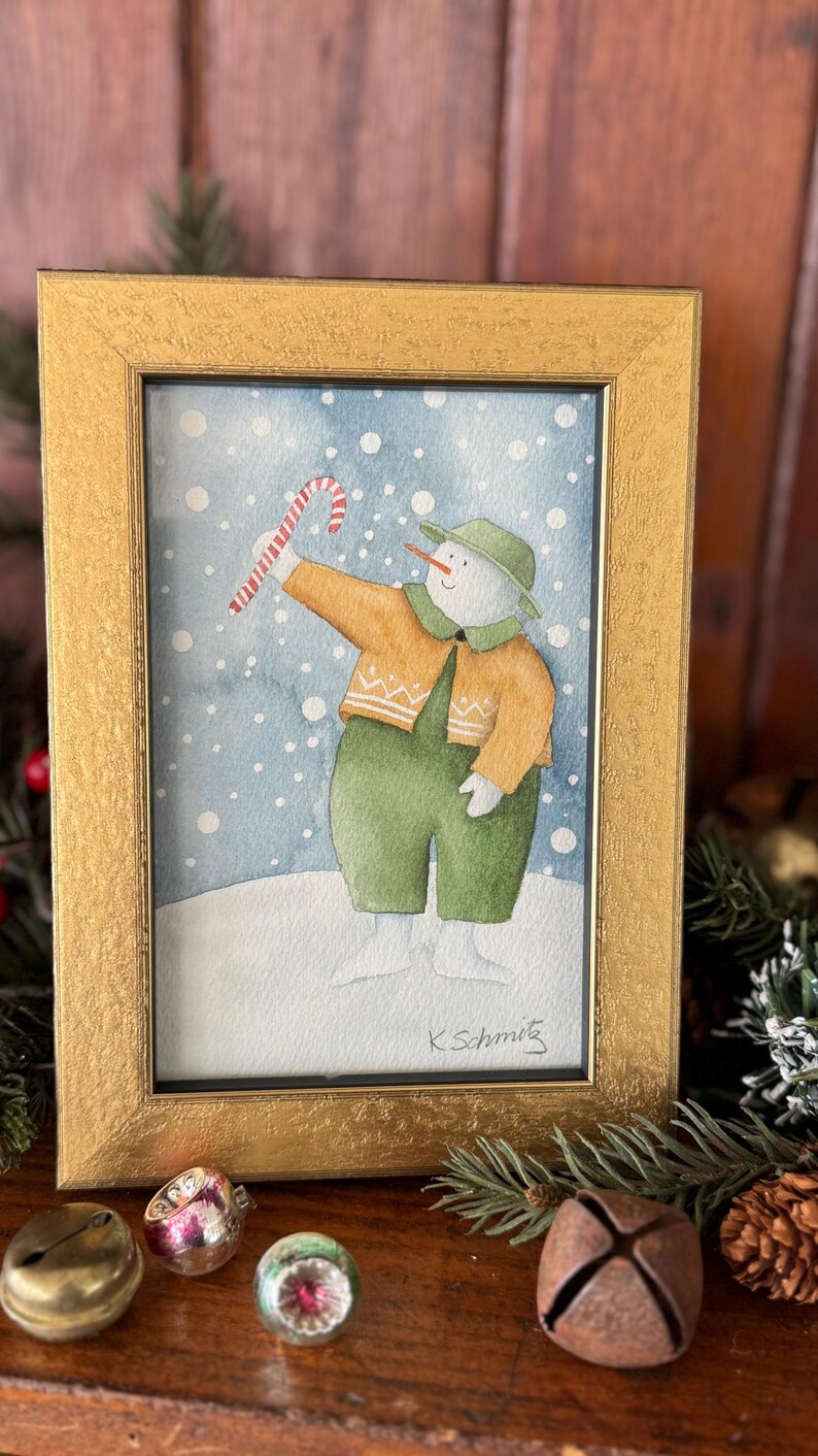 K&ouml;nnte beinhalten: Aquarellmalerei eines Schneemanns in einem goldenen Rahmen. Der Schneemann tr&auml;gt einen gr&uuml;nen Hut, eine Latzhose und einen orangefarbenen Pullover und h&auml;lt eine Zuckerstange. Der Hintergrund ist eine verschneite blaue Szene. Das Gem&auml;lde ist signiert "K. Schmitz".