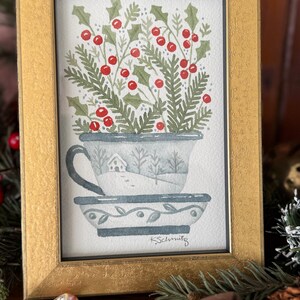 K&ouml;nnte beinhalten: Ein gerahmtes Aquarellgem&auml;lde einer Teetasse, gef&uuml;llt mit Stechpalmen und Beeren. Die Teetasse ist mit einer Winterszene verziert. Der Rahmen ist goldfarben. Das Gem&auml;lde ist mit "K. Schmitz" signiert.