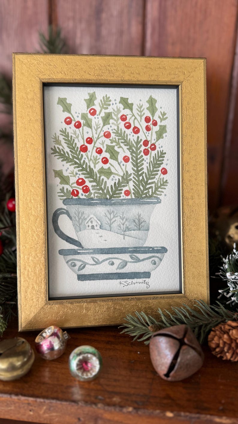 K&ouml;nnte beinhalten: Ein gerahmtes Aquarellgem&auml;lde einer Teetasse, gef&uuml;llt mit Stechpalmen und Beeren. Die Teetasse ist mit einer Winterszene verziert. Der Rahmen ist goldfarben. Das Gem&auml;lde ist mit "K. Schmitz" signiert.
