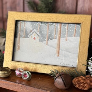 K&ouml;nnte beinhalten: Ein gerahmtes Aquarellgem&auml;lde zeigt eine verschneite Winterszene mit einem kleinen Haus und B&auml;umen. Das Gem&auml;lde befindet sich in einem goldenen Rahmen und ist mit "K. Schmitz" signiert. Dekorative Elemente wie Glocken und Ornamente deuten auf ein Feiertags-Thema hin.