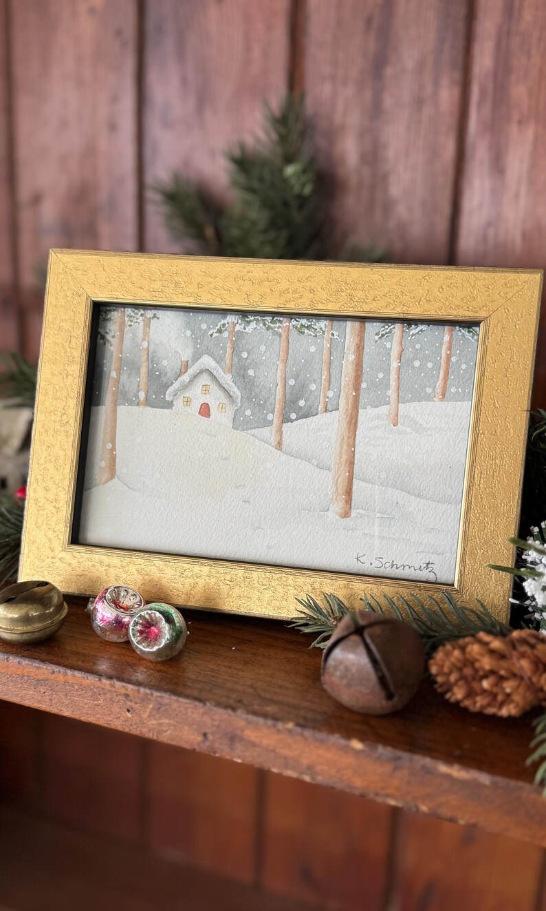 K&ouml;nnte beinhalten: Ein gerahmtes Aquarellgem&auml;lde zeigt eine verschneite Winterszene mit einem kleinen Haus und B&auml;umen. Das Gem&auml;lde befindet sich in einem goldenen Rahmen und ist mit "K. Schmitz" signiert. Dekorative Elemente wie Glocken und Ornamente deuten auf ein Feiertags-Thema hin.