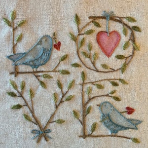 Pode incluir: Bordado com dois pássaros azuis empoleirados em galhos com pequenos corações vermelhos. Os galhos formam as letras "LOVE" com um coração vermelho maior pendurado no "O".